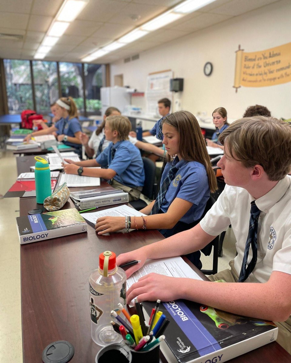 Uniformed students at Coram Deo Academy work through a biology lesson at long classroom tables, textbooks and papers open in front of them.