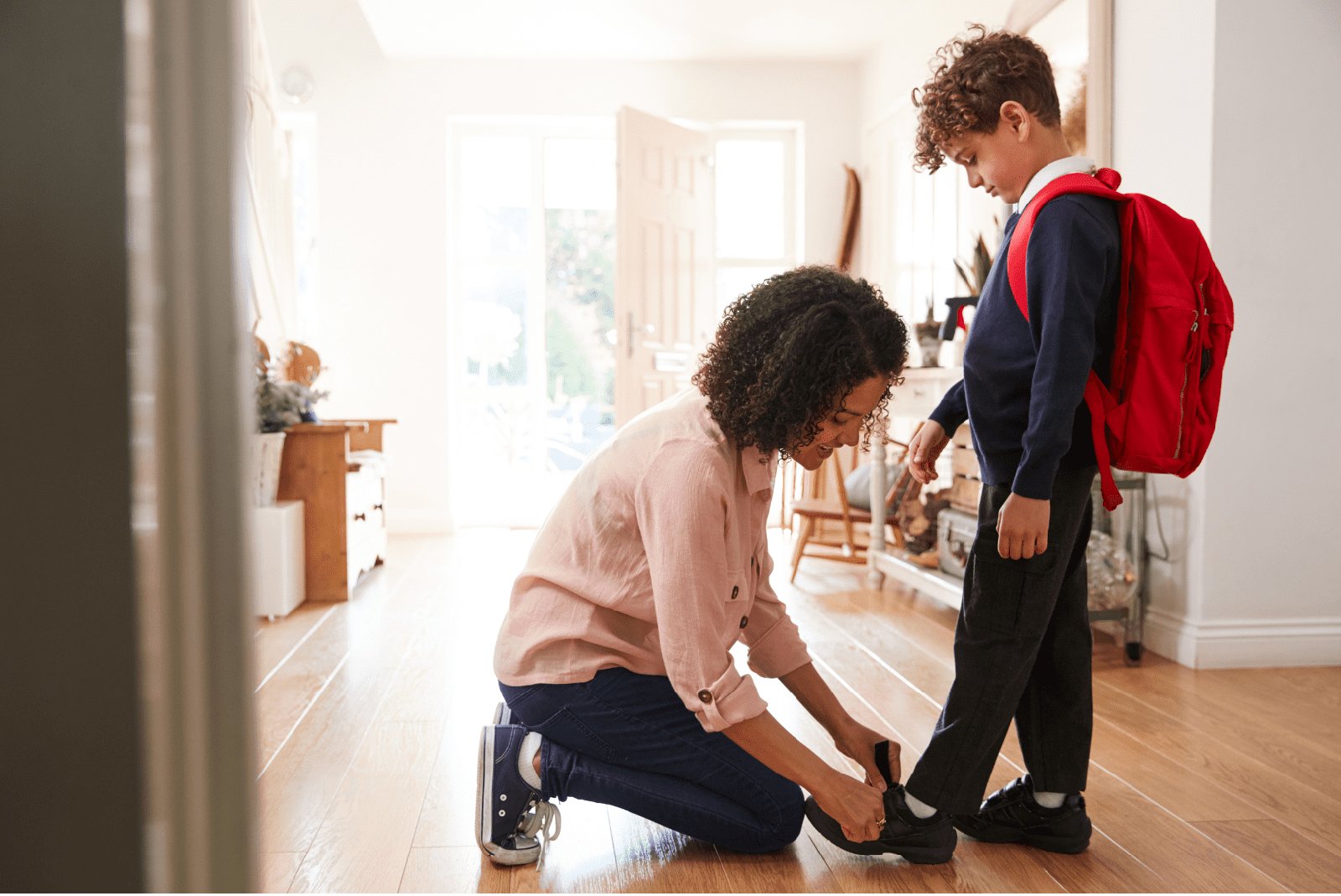 A mother kneels in the front hallway to help her son fasten his shoes before school, his red backpack already on his shoulders.