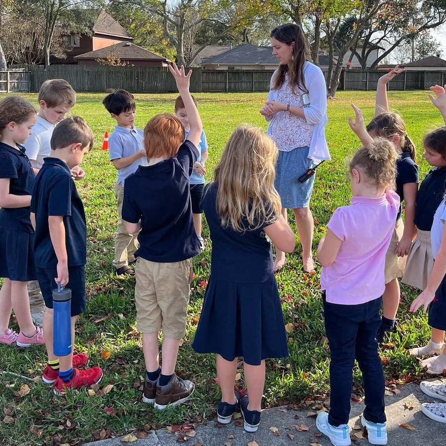 A teacher gathers a circle of uniformed elementary students on a member-school lawn as several raise their hands
